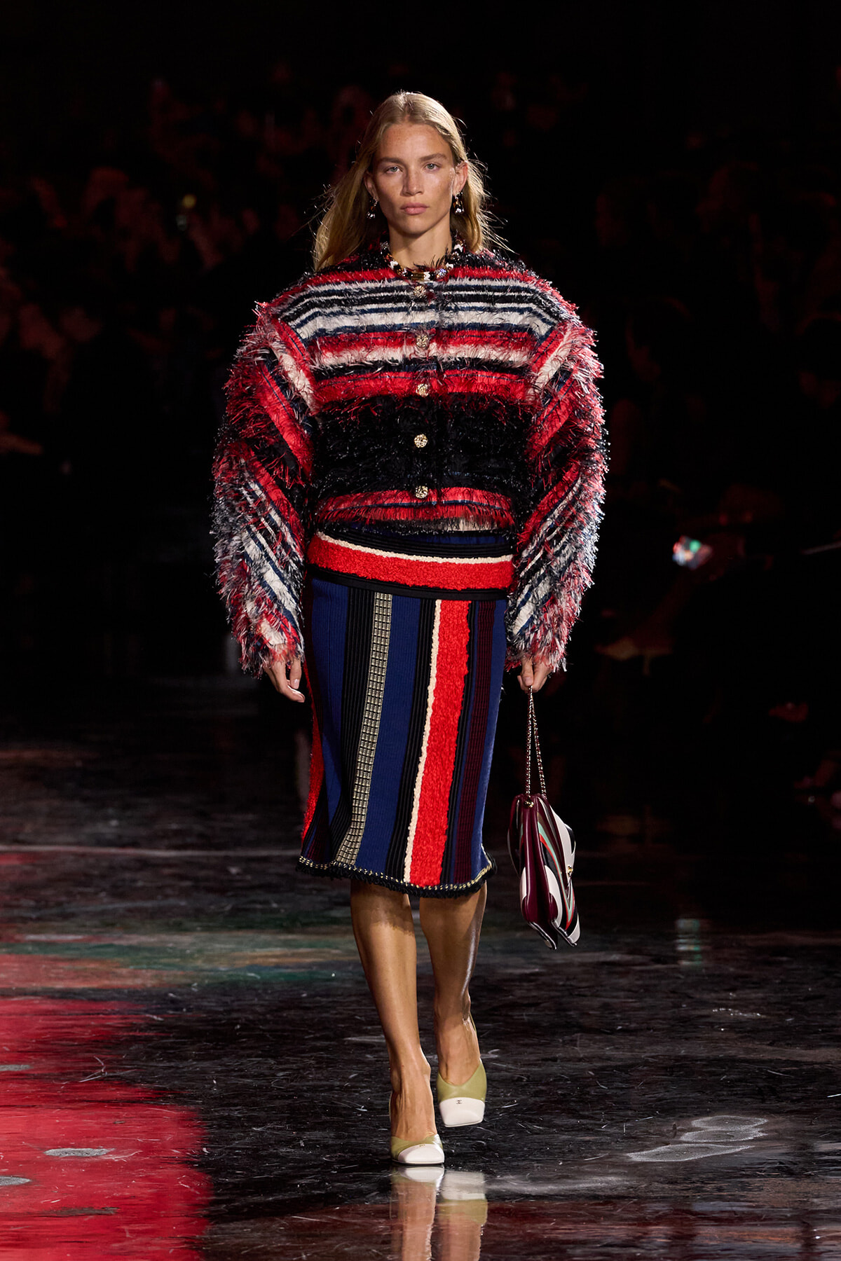 Model walking a fashion show runway in a multicolored, fringed knit sweater and matching striped skirt, carrying a striped handbag.