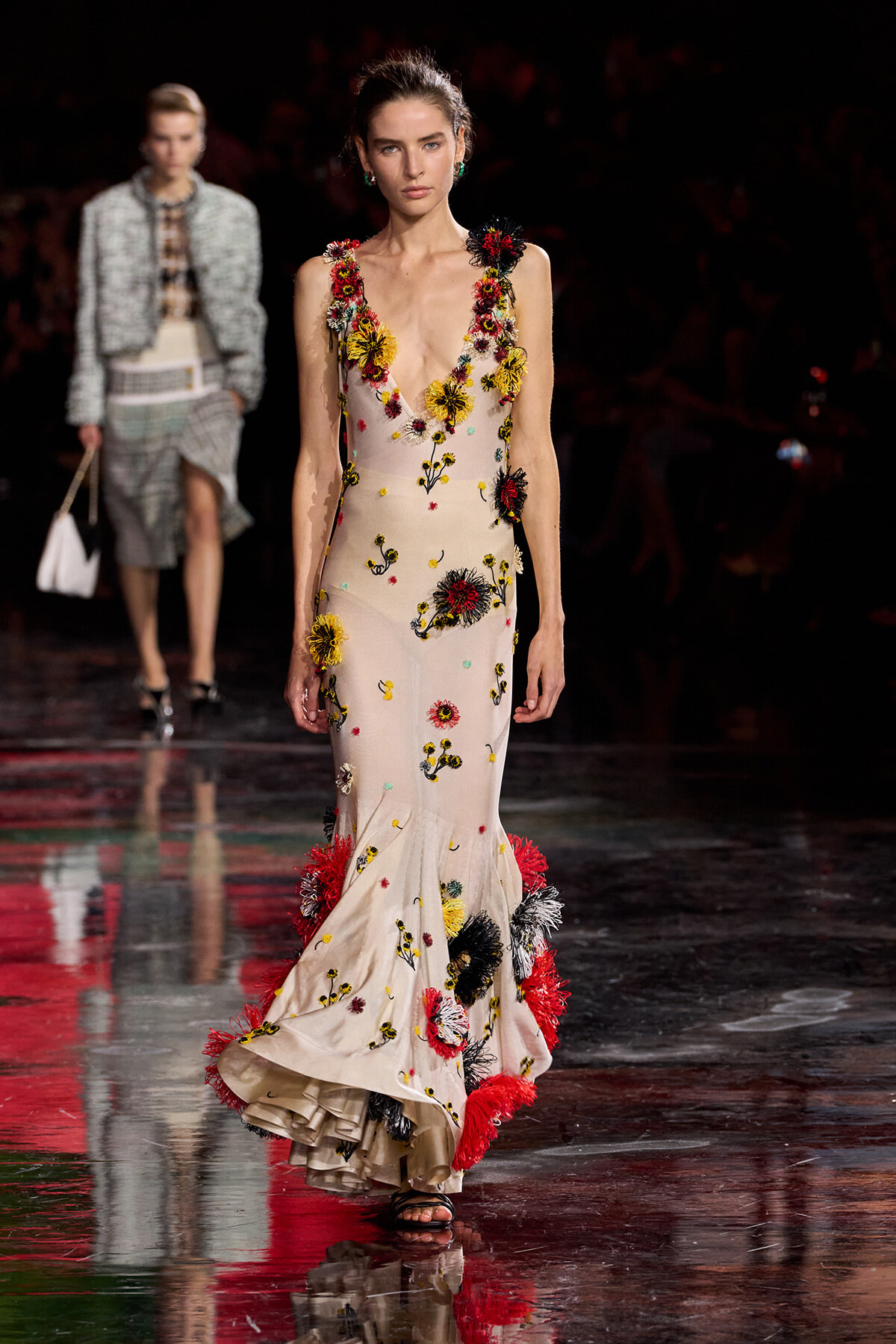 Runway model wearing a cream, floor-length gown densely embroidered with colorful 3D flowers along the neckline and hem, walking on a glossy catwalk.