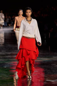 Model walking a runway in a white blouse and high-low red ruffled skirt with a tan belt and chunky necklace, holding a dark clutch.