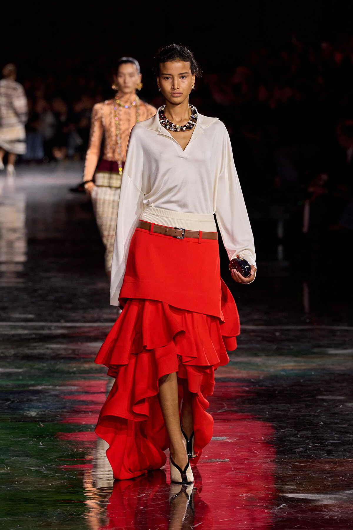 Model walking a runway in a white blouse and high-low red ruffled skirt with a tan belt and chunky necklace, holding a dark clutch.