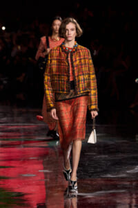 Model walking the runway in a yellow-orange plaid jacket over a red plaid skirt, paired with a bold beaded necklace and a small white handbag.