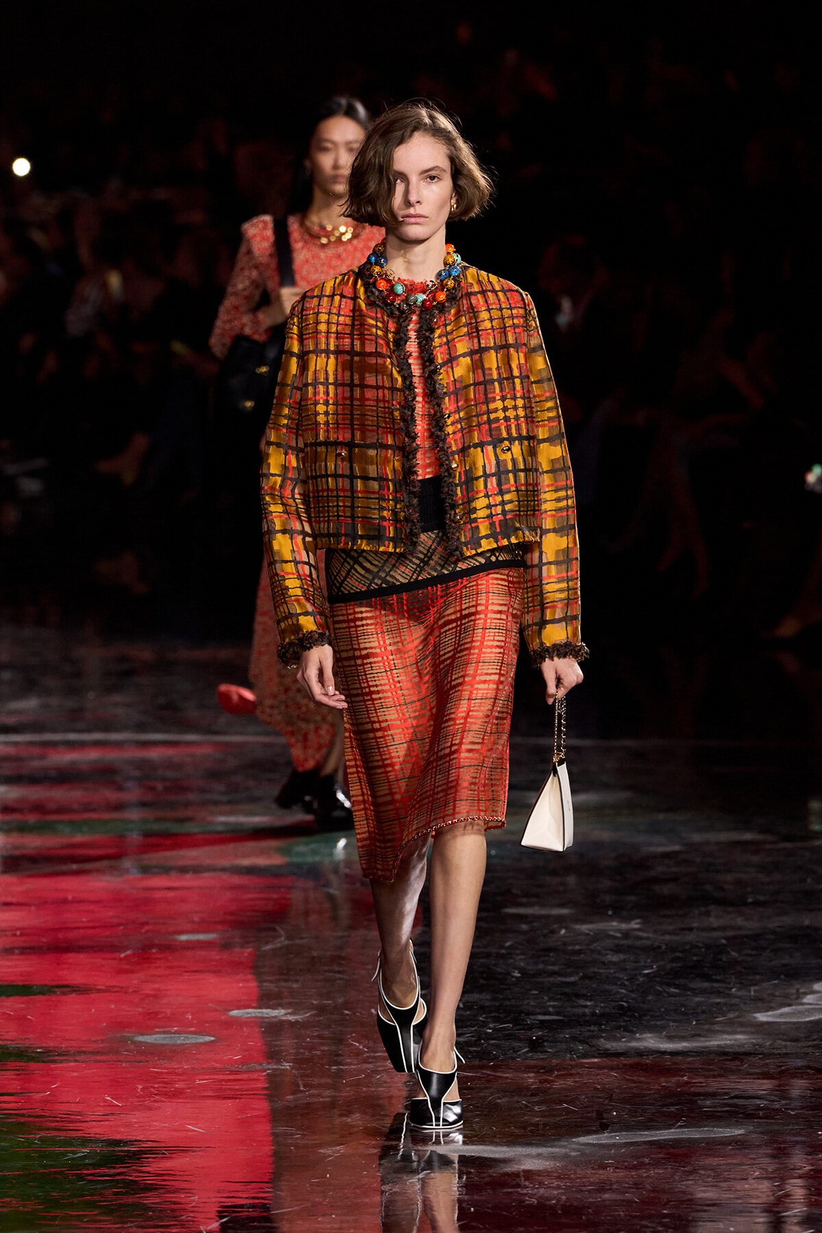Model walking the runway in a yellow-orange plaid jacket over a red plaid skirt, paired with a bold beaded necklace and a small white handbag.