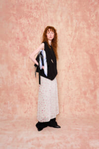 Red-haired woman in a black sleeveless top and long white skirt with pink speckles, standing against a peach textured wall while holding a bag with a blue scarf.