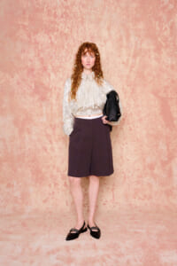 Woman with curly red hair stands against a pink textured wall, wearing a metallic beige blouse, dark shorts, black flats, and carrying a black bag.