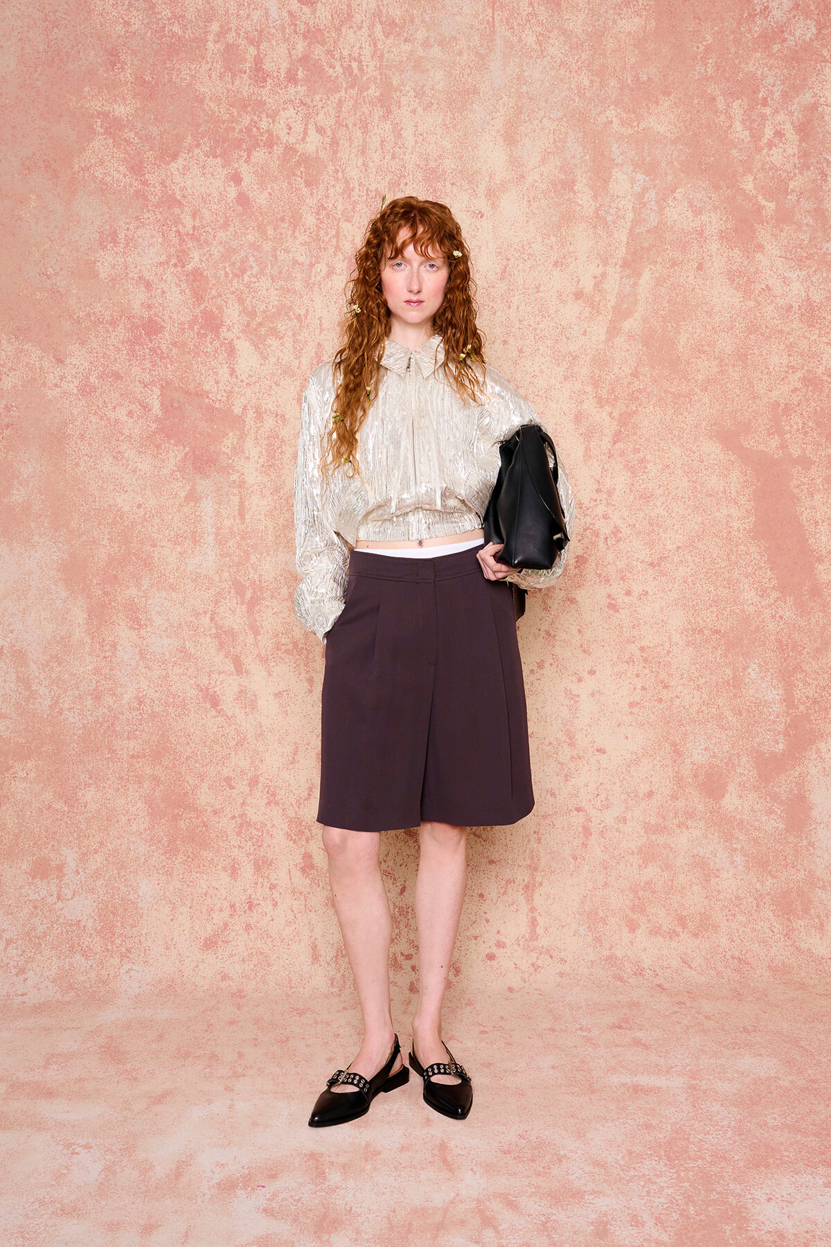 Woman with curly red hair stands against a pink textured wall, wearing a metallic beige blouse, dark shorts, black flats, and carrying a black bag.
