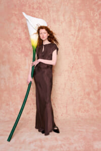 Woman in a brown, full-length dress stands against a pink textured backdrop, holding a giant white calla lily with a green stem.