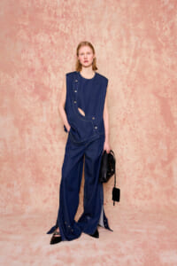 Model in a sleeveless dark denim top with a cutout, paired with wide-leg denim pants, standing against a peach textured backdrop while holding a black bag.