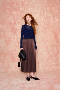 Full-length portrait of a red-haired woman in a navy top and brown long skirt, holding a black handbag, standing against a peach textured backdrop.