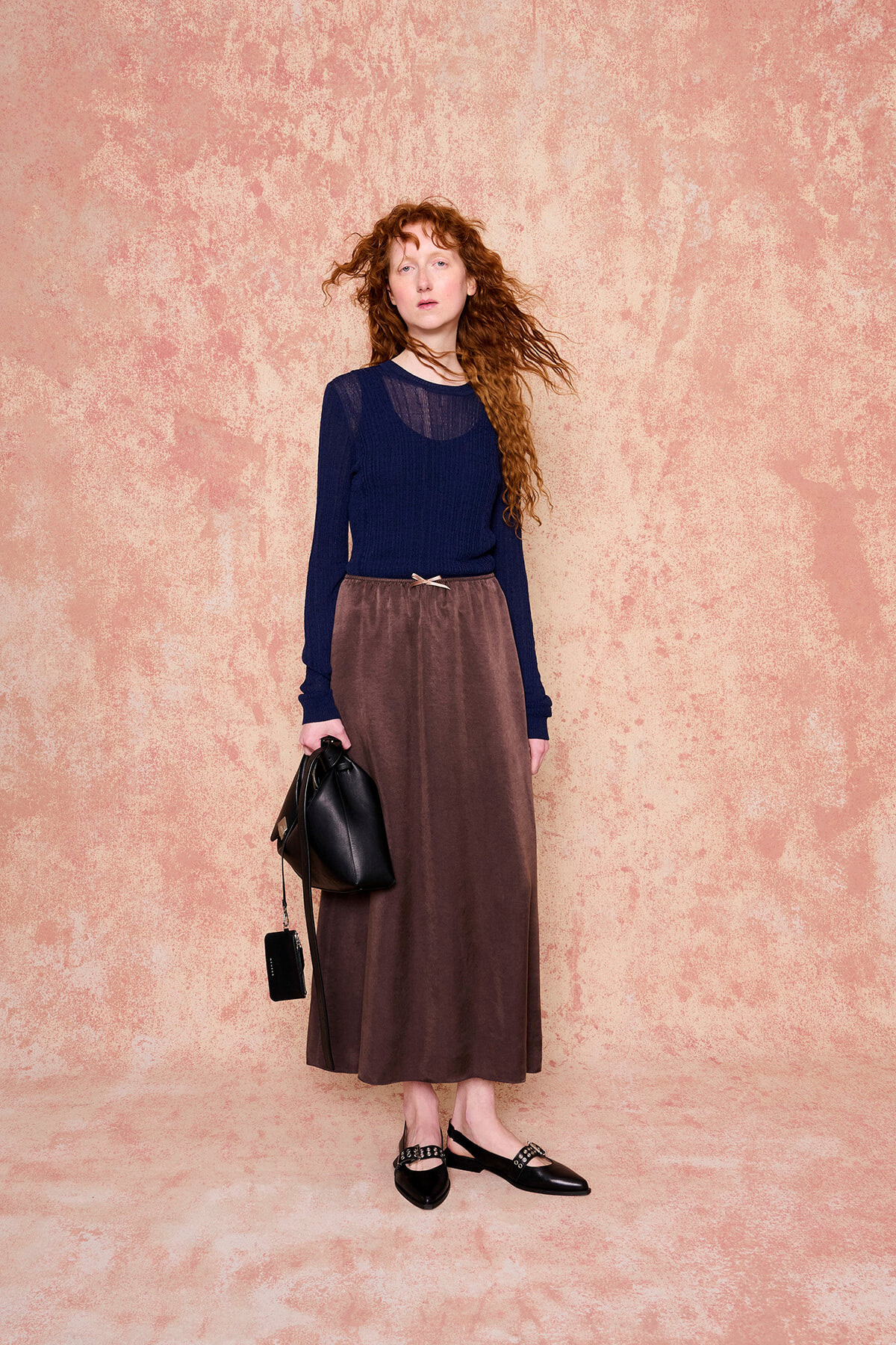 Full-length portrait of a red-haired woman in a navy top and brown long skirt, holding a black handbag, standing against a peach textured backdrop.