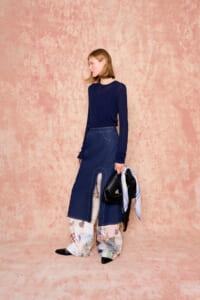 Woman wearing a navy knit top and a dark denim skirt with a front slit revealing patterned pants, holding a black handbag and striped scarf against a peach backdrop.
