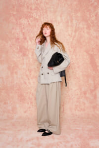 Woman with wavy red hair in a beige oversized suit, carrying a black handbag, standing against a pink textured backdrop.