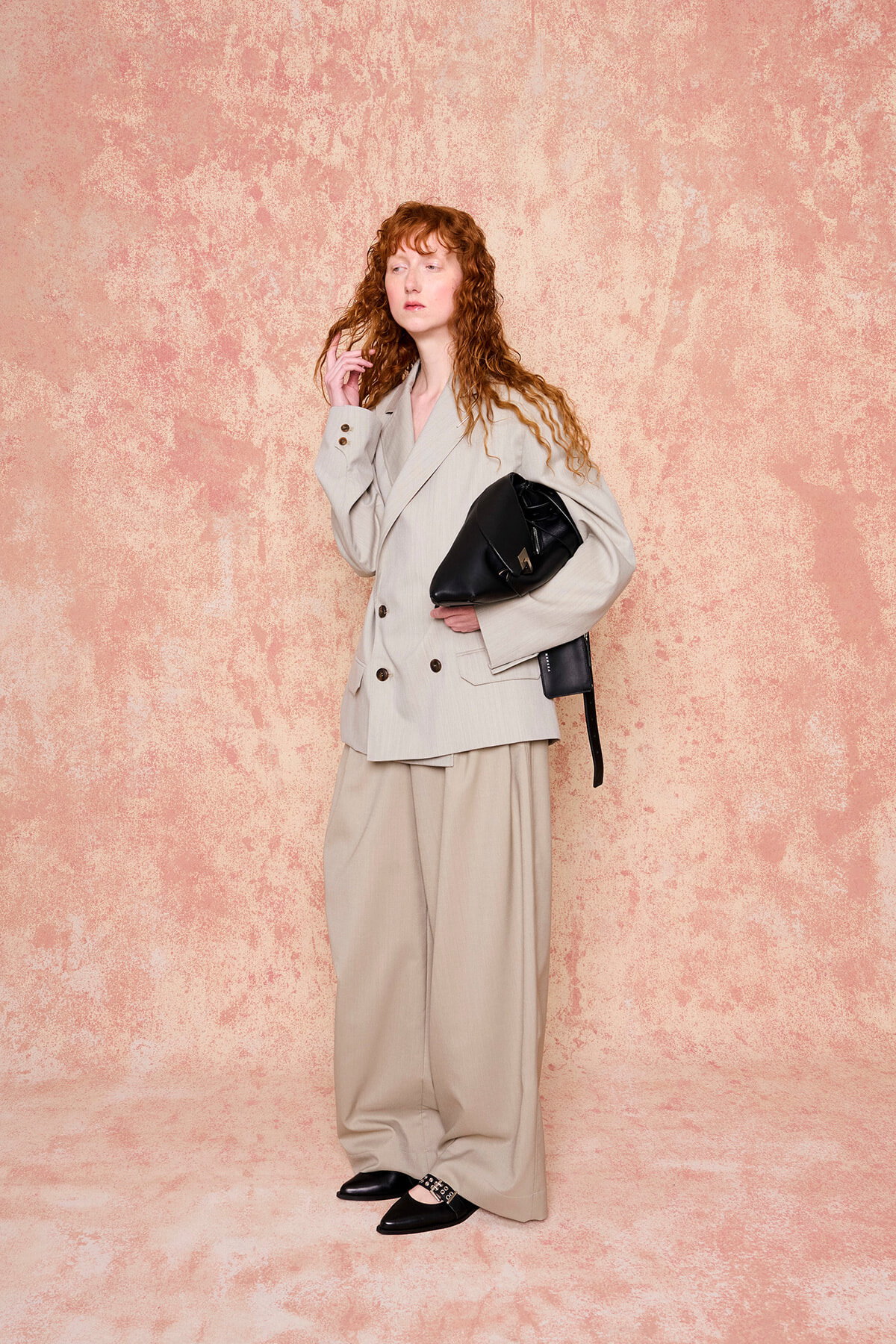 Woman with wavy red hair in a beige oversized suit, carrying a black handbag, standing against a pink textured backdrop.