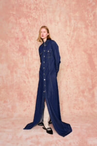 Model wearing a full-length dark denim button-front dress with chest pockets, standing against a pink textured backdrop.