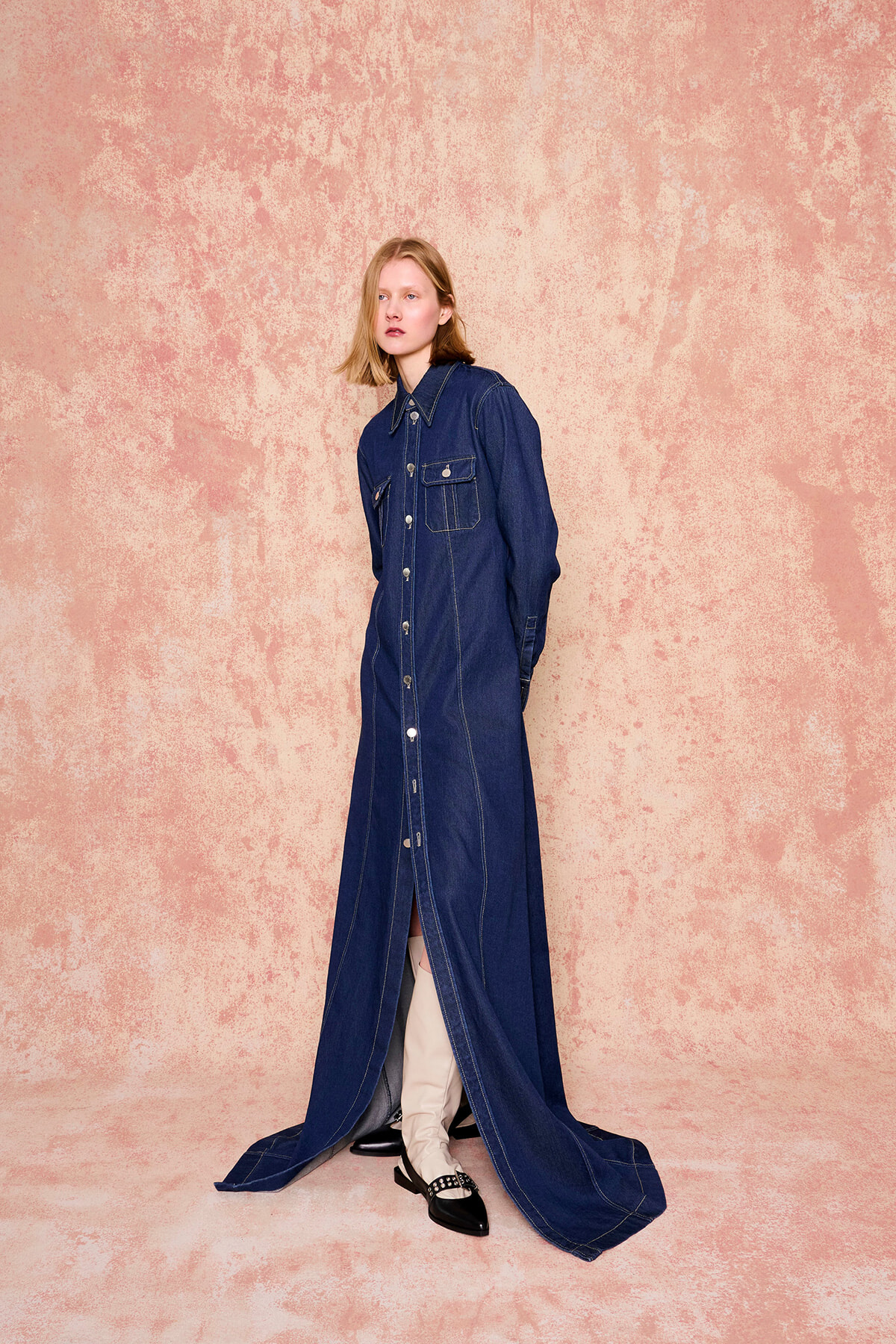 Model wearing a full-length dark denim button-front dress with chest pockets, standing against a pink textured backdrop.
