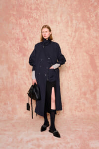 Model in a navy long coat with diagonal front snaps, gray sleeve cuffs, and a high slit, carrying a black handbag against a pink textured backdrop.