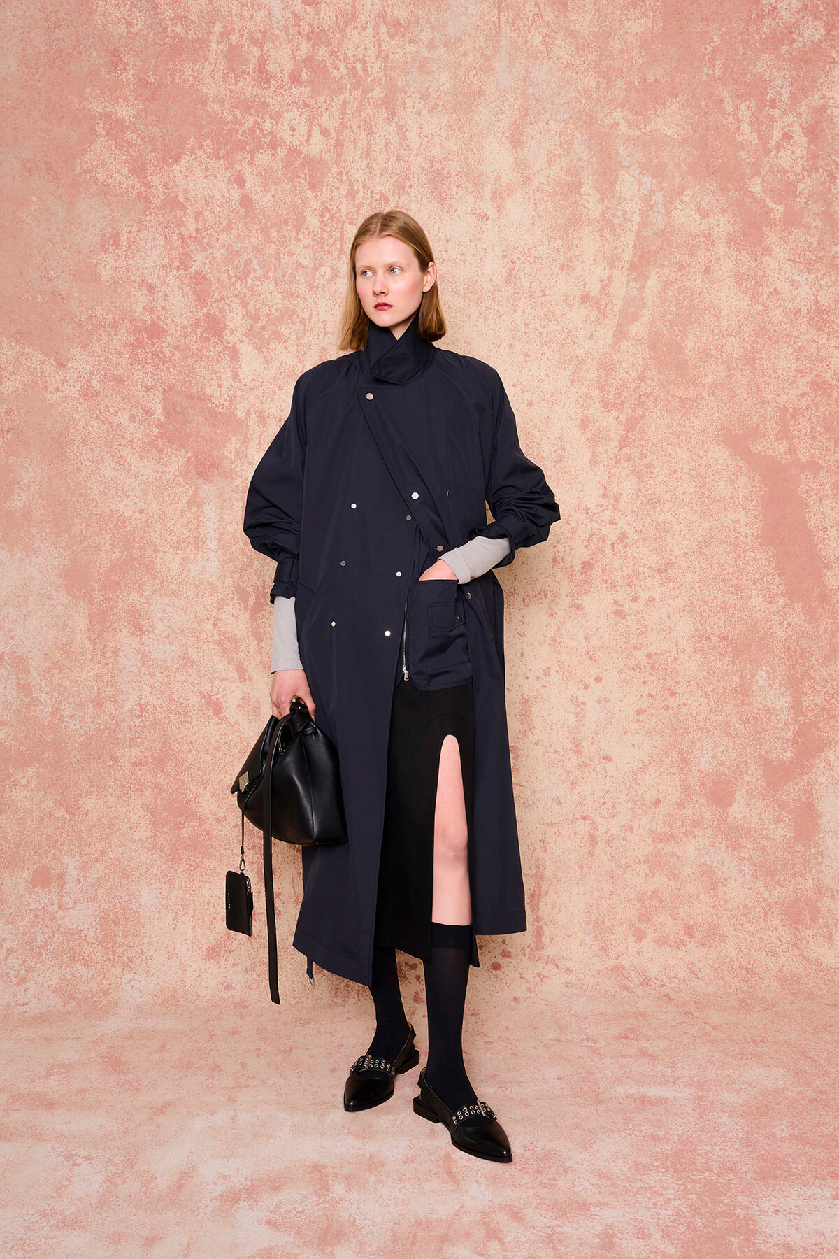 Model in a navy long coat with diagonal front snaps, gray sleeve cuffs, and a high slit, carrying a black handbag against a pink textured backdrop.