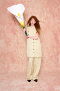 Woman with long red hair in beige outfit holding a large white calla lily against a pink textured background.