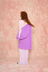 Woman with curly red hair seen from the back, wearing a lavender dress layered over a light pink top against a textured pink wall.