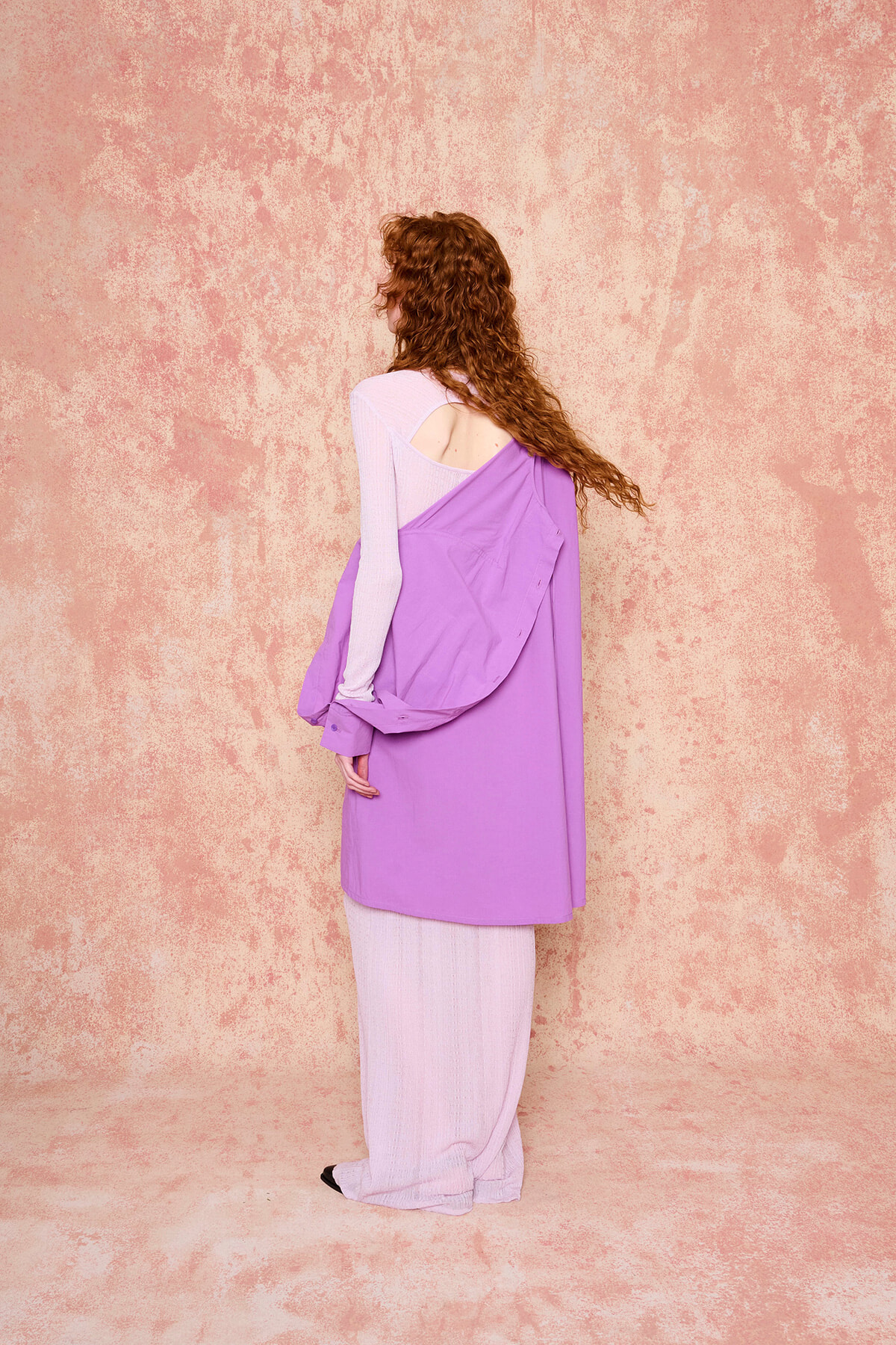 Woman with curly red hair seen from the back, wearing a lavender dress layered over a light pink top against a textured pink wall.
