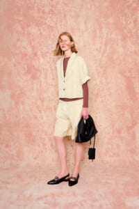 Model wearing a cream blazer vest over a maroon top and matching shorts, holding a black handbag in a studio with a pink textured backdrop.