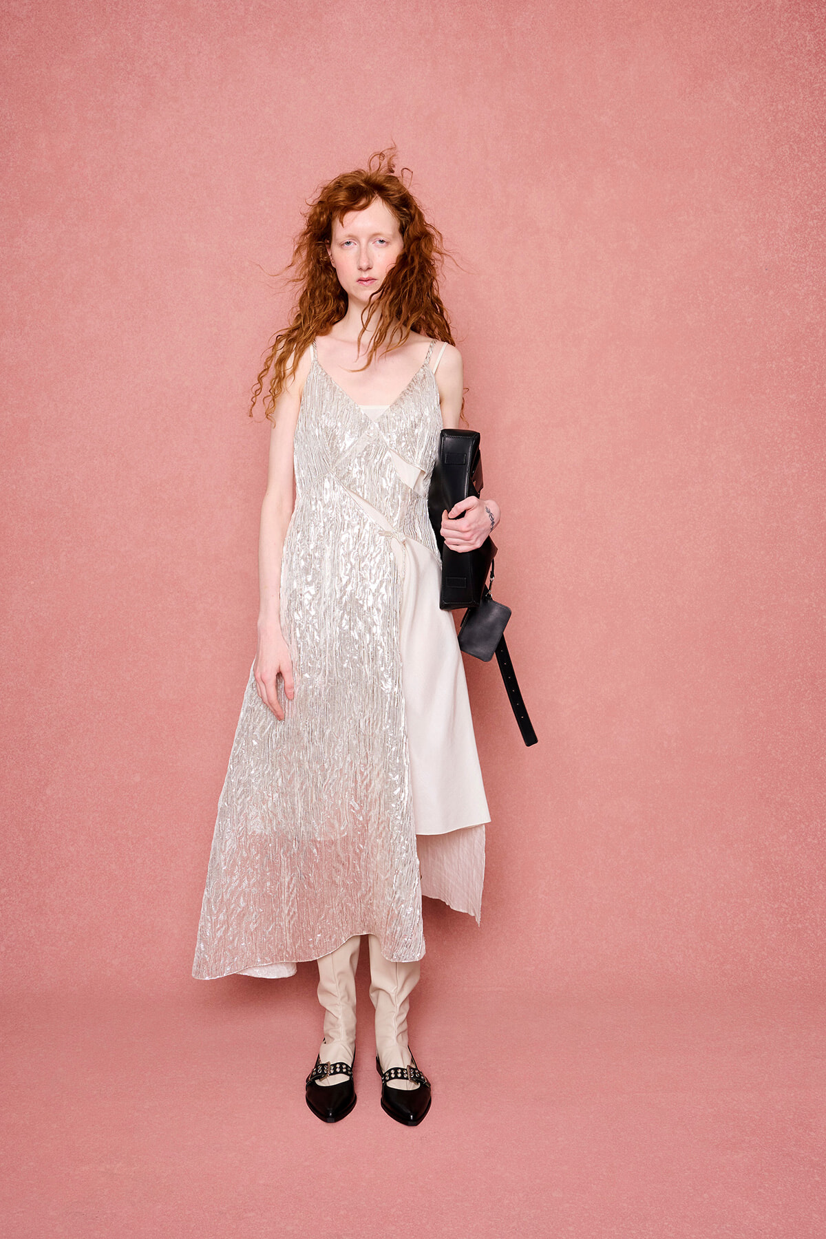 Woman standing against a pink backdrop in a light metallic dress with layered panels, beige boots, and a black handbag held at the side.