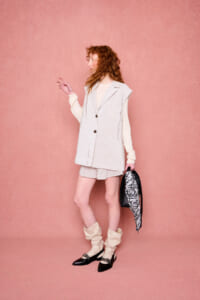 Full-body fashion shot: person with wavy red hair wearing a beige pinstripe sleeveless blazer over a cream sweater and matching shorts, holding a black-and-white patterned scarf bag against a pink backdrop.