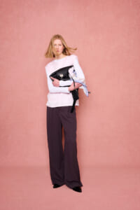 Model stands against pink backdrop, wearing a white long-sleeve top and wide-leg dark pants, clutching a black handbag with a scarf draped over it.