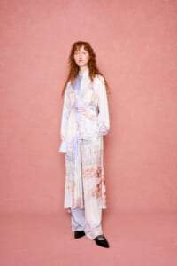 Woman standing against a pink textured backdrop in a pastel wrap dress with handwritten script prints; hands at her sides, neutral expression, black shoes visible.