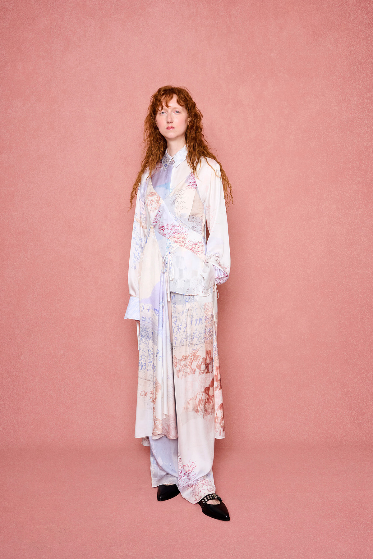 Woman standing against a pink textured backdrop in a pastel wrap dress with handwritten script prints; hands at her sides, neutral expression, black shoes visible.