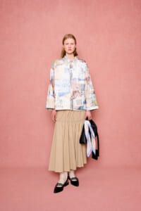 Model in pastel patchwork blouse with a gathered beige midi skirt, carrying a black handbag with a scarf, against a pink backdrop.