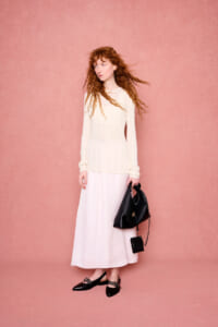 Woman with long curly red hair in a cream knit top and white skirt stands against a pink backdrop, holding a black handbag.