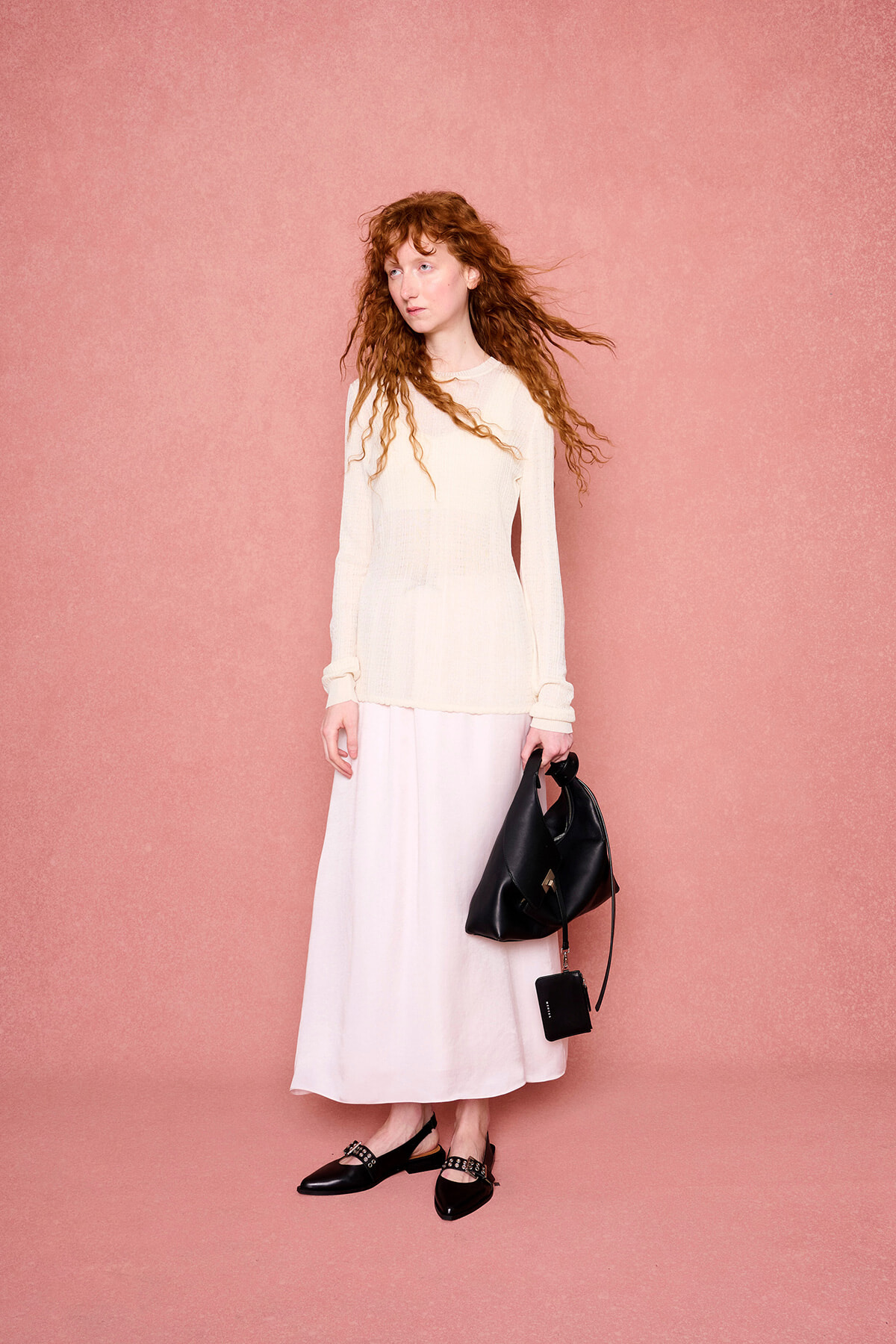 Woman with long curly red hair in a cream knit top and white skirt stands against a pink backdrop, holding a black handbag.