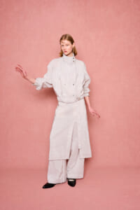 Woman wearing a light striped high-neck coat with wide-leg pants, posing against a pink textured backdrop.