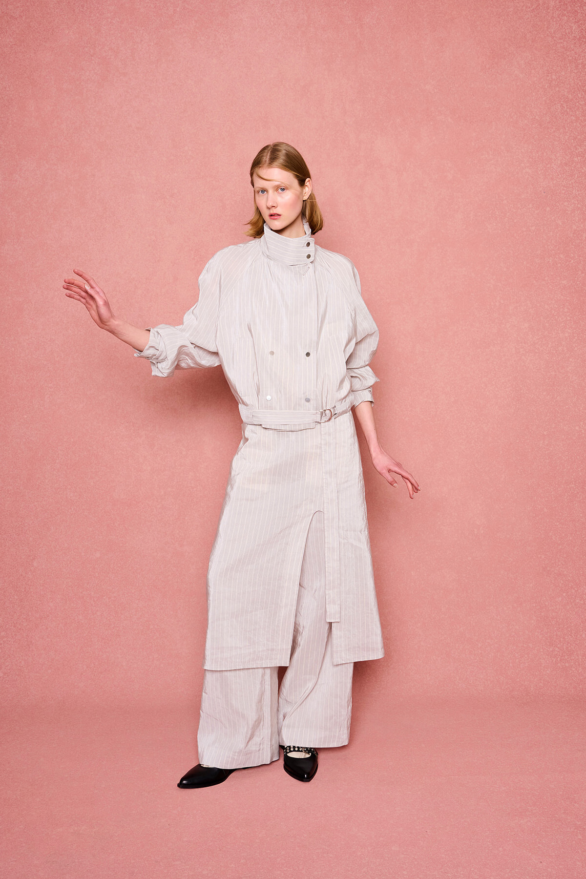 Woman wearing a light striped high-neck coat with wide-leg pants, posing against a pink textured backdrop.