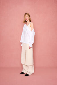 Woman in a loose white shirt over a beige dress, standing against a pink textured backdrop; minimalist fashion look with black pointed shoes nearby.