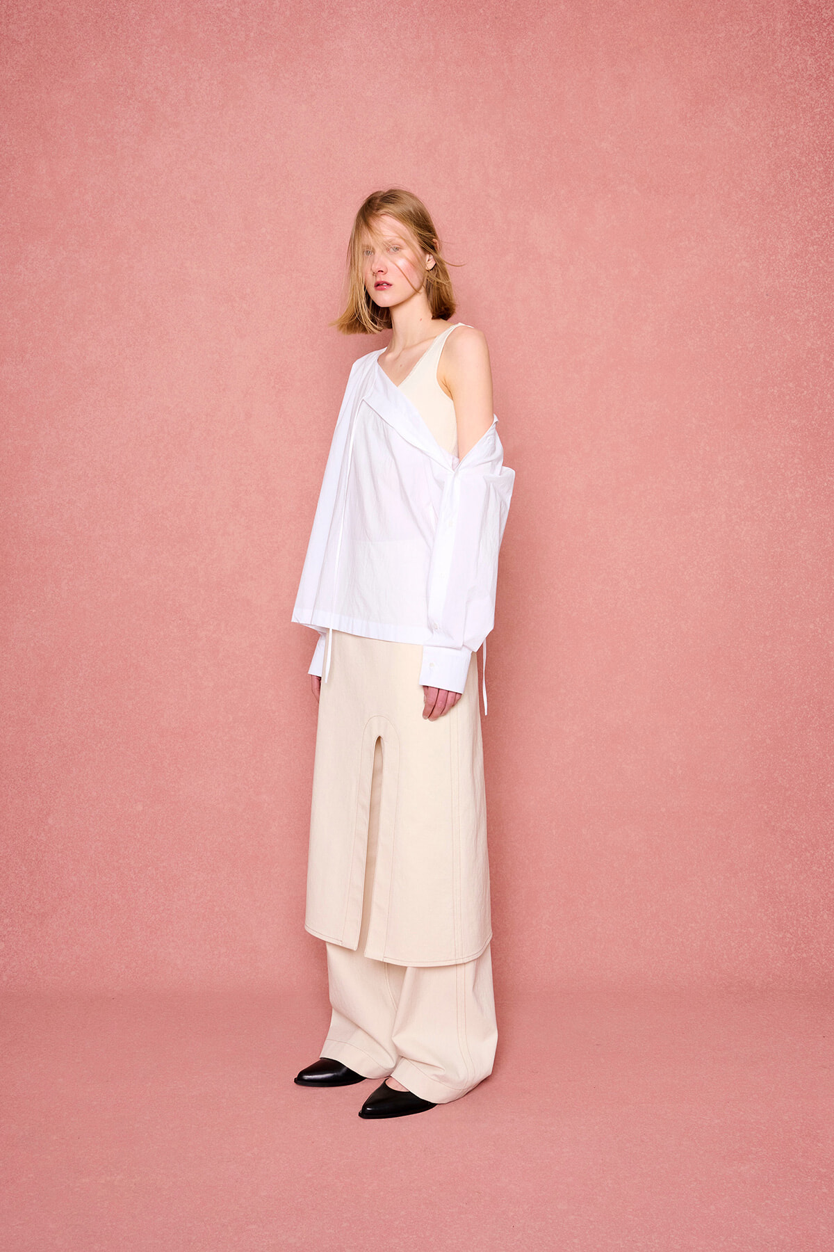 Woman in a loose white shirt over a beige dress, standing against a pink textured backdrop; minimalist fashion look with black pointed shoes nearby.