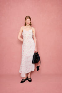 Woman wearing a light pink floral slip dress with one-shoulder strap, standing against a pink backdrop, holding a black handbag.