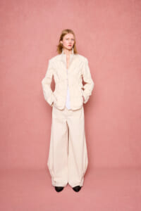 Model in oversized cream suit with a white shirt, standing against a pink backdrop, hands in pockets, neutral expression.