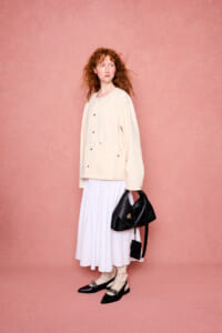 Woman with curly red hair stands against a pink backdrop in a cream jacket and white skirt, holding a black handbag.