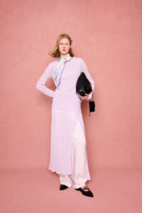 Model in pastel lavender knit top and skirt over white pants, with a light scarf, holding a black handbag against a pink backdrop.