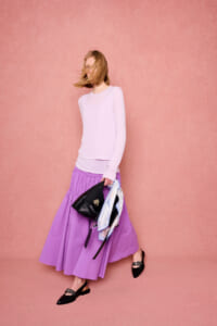 Woman in a light lavender knit top and a long purple skirt walks against a pink textured wall, carrying a black handbag and a scarf.