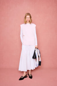 Model stands against a pink backdrop in a pale pink tunic over a white long-sleeve top and a white tiered midi skirt, holding a black bag and scarf.