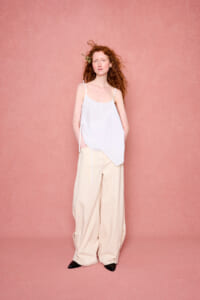 Woman with long red hair standing against a pink wall, wearing a white asymmetrical top and beige wide-leg pants.