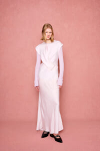 Model wearing a pale pink satin gown with a draped bodice over a pink knit top, standing against a pink textured backdrop.