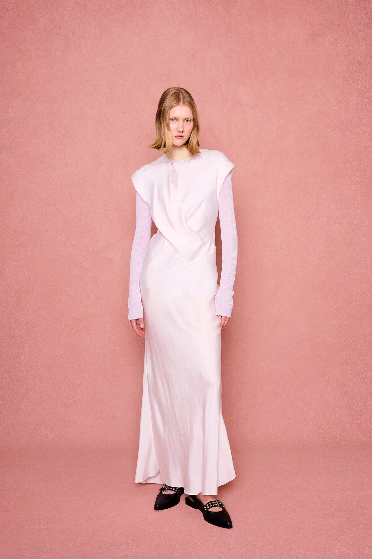 Model wearing a pale pink satin gown with a draped bodice over a pink knit top, standing against a pink textured backdrop.