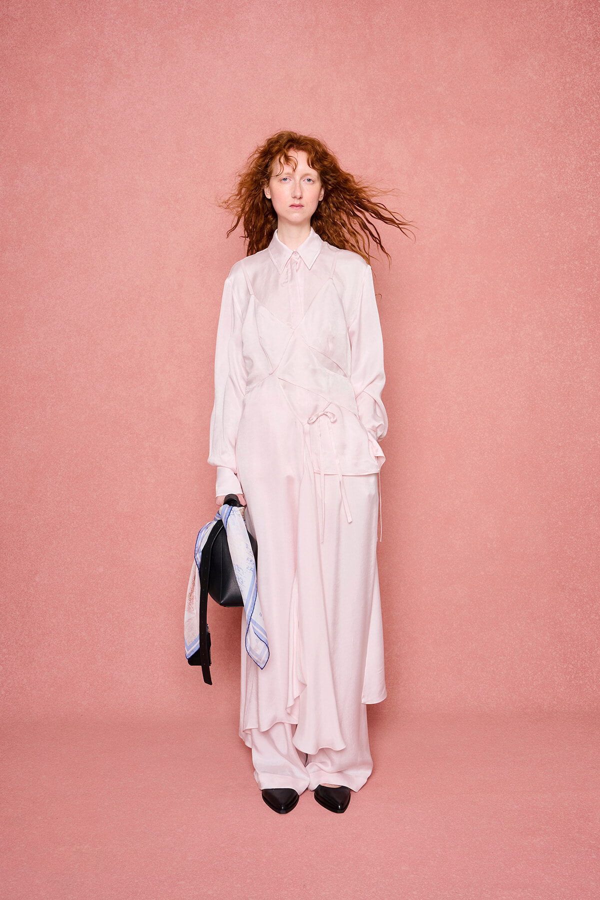 Model in pale pink satin outfit standing against a pink backdrop, holding a black bag with a blue scarf拢