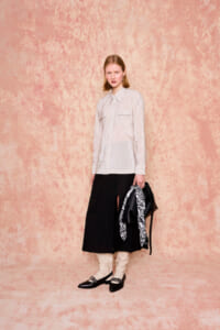 Woman standing in a peach textured studio backdrop wearing a light striped button-down shirt, a black midi skirt with a front slit, cream boots, and holding a black bag with a black-and-white patterned scarf.