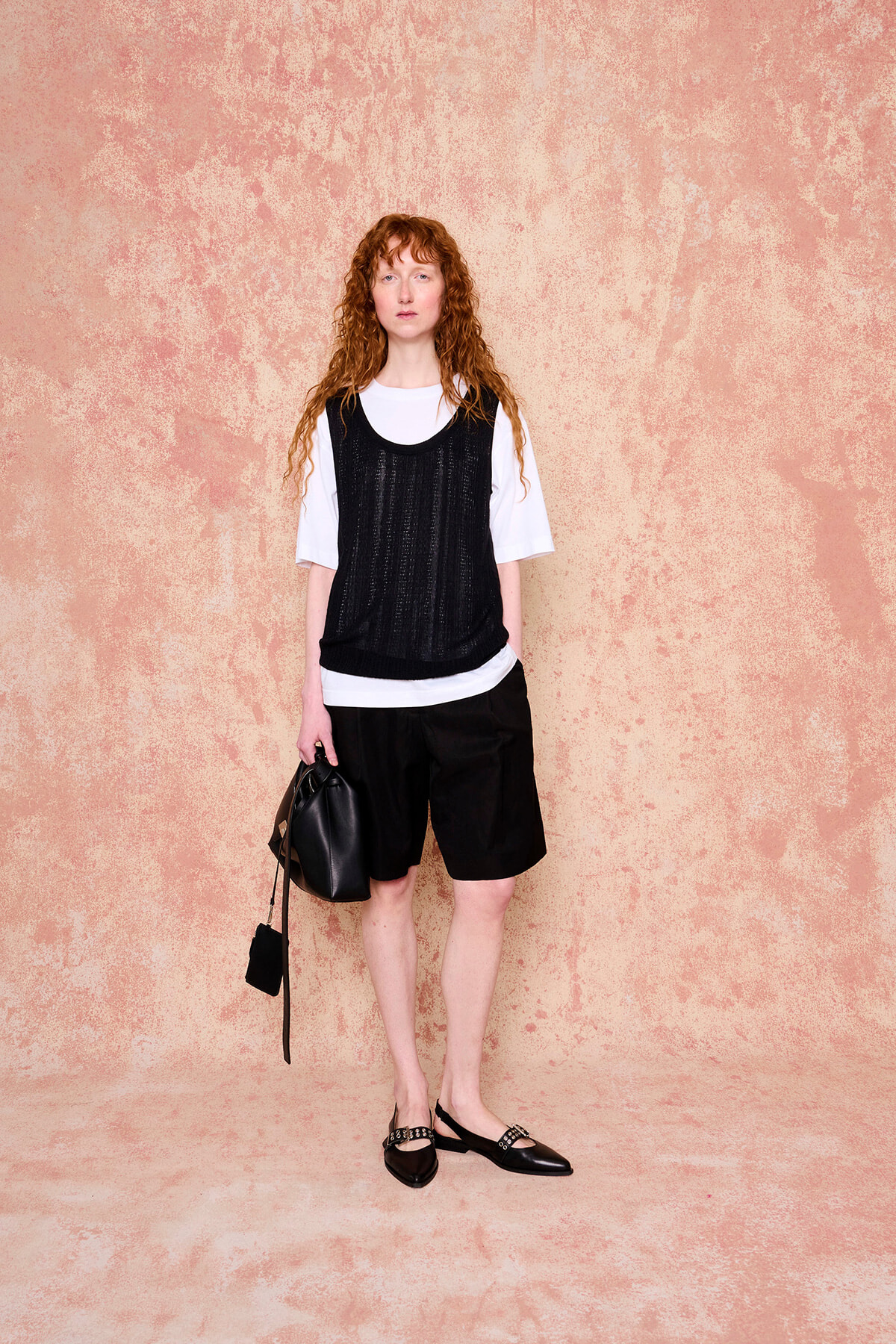 Full-body fashion portrait of a red-haired woman in a white T-shirt, black mesh vest, black shorts, and black flats, holding a black bag against a peach wall.