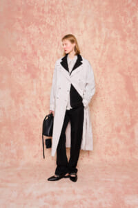 Person wearing a pale pinstriped trench coat over a gray shirt and black pants, holding a black handbag against a pink textured backdrop.