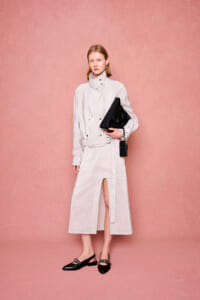 Female model in a light striped trench dress with high collar, holding a black clutch against a pink backdrop.